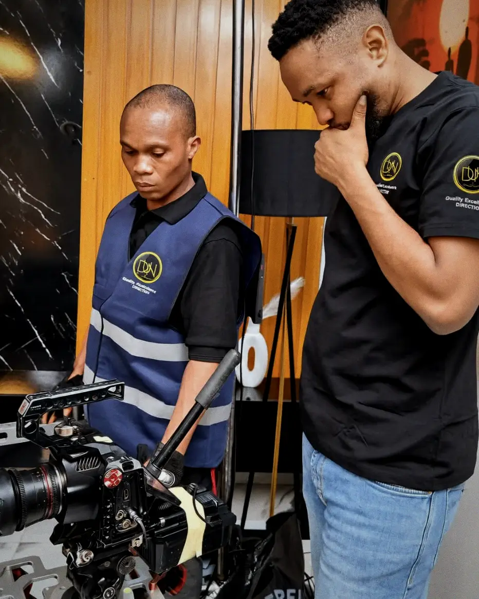 Two DUN crew members at a cinema camera rig against a warm timber wall, one operating, one considering the shot.